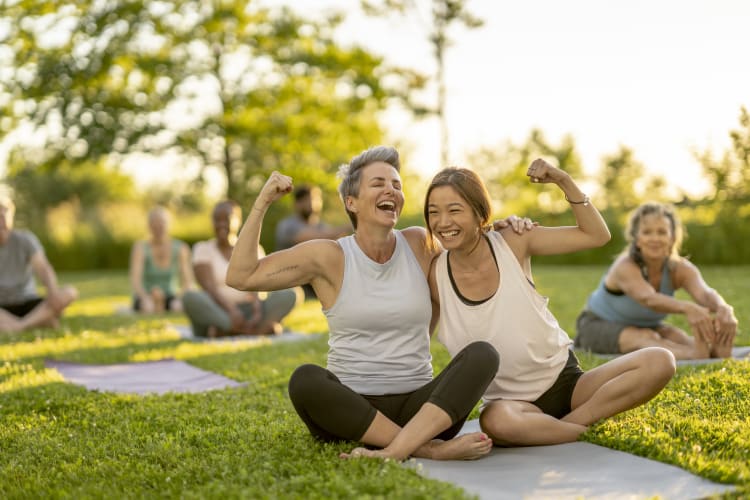Two laughing woman sit on a yoga mat outside as they lean in closely for a hug and flex their muscles.
